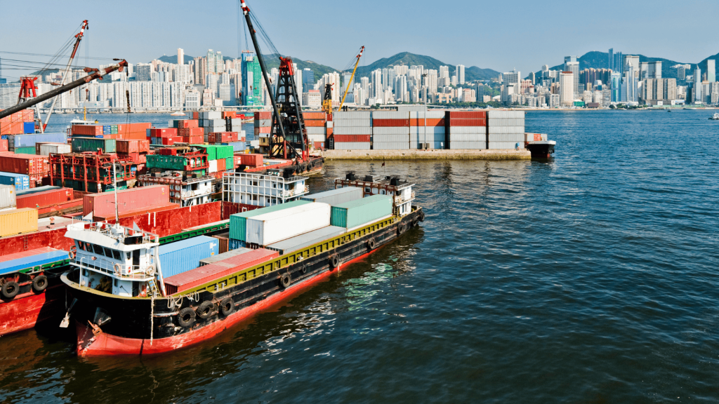 Busy Asian port scene with container barges and cranes in the foreground and a dense city skyline in the background.