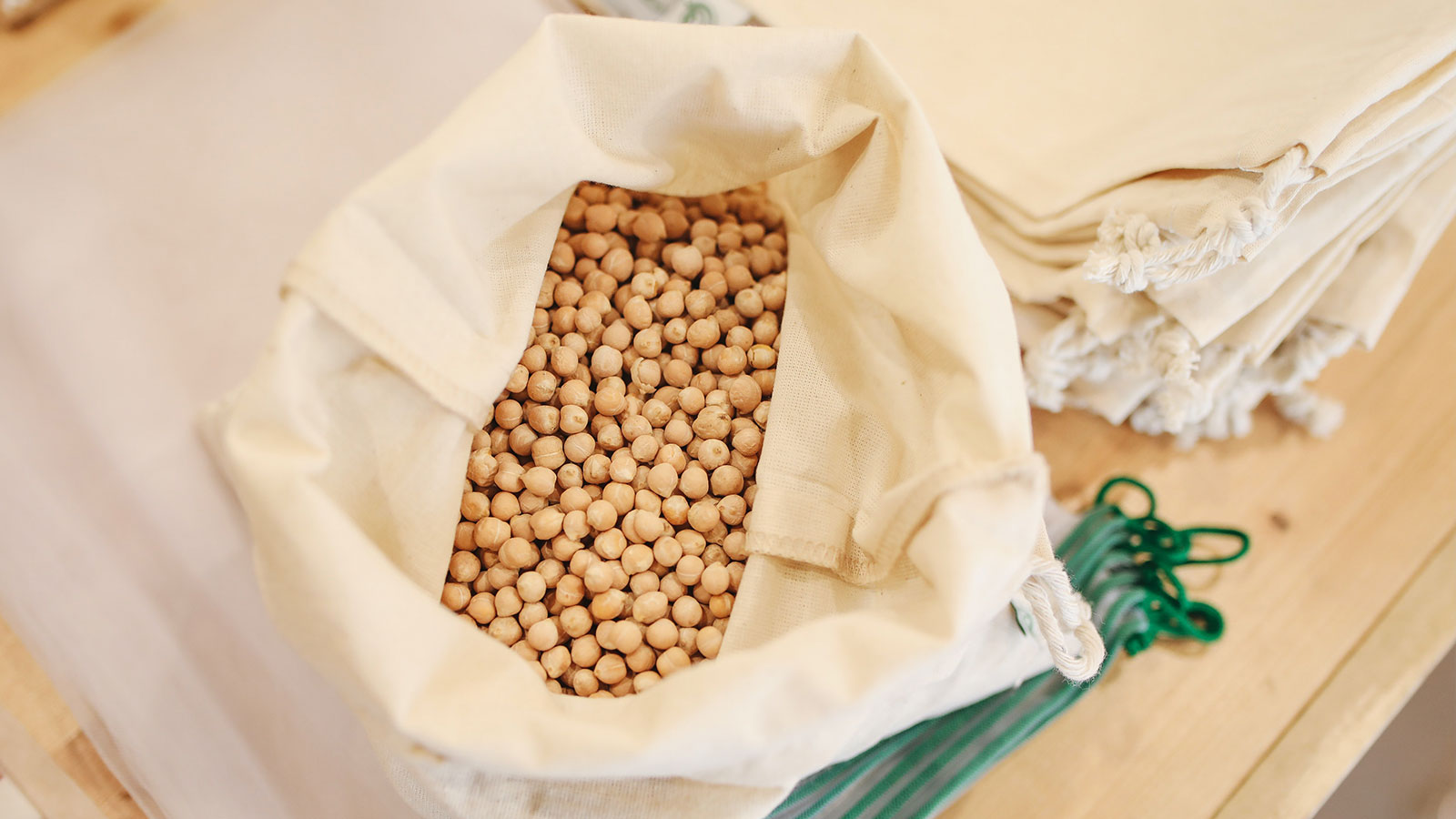 Overhead view of a tan cloth bag filled with dry, raw chickpeas (garbanzo beans) on a wooden table next to reusable cloth sacks.
